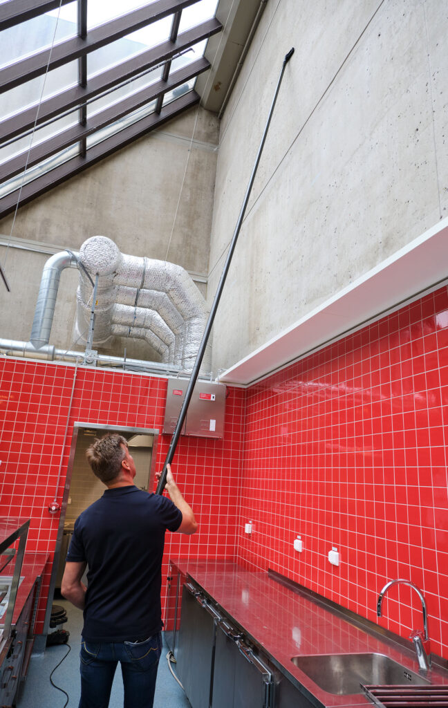Operator using a ground-based high-reach pole to clean high-level fittings in a red-tiled commercial kitchen