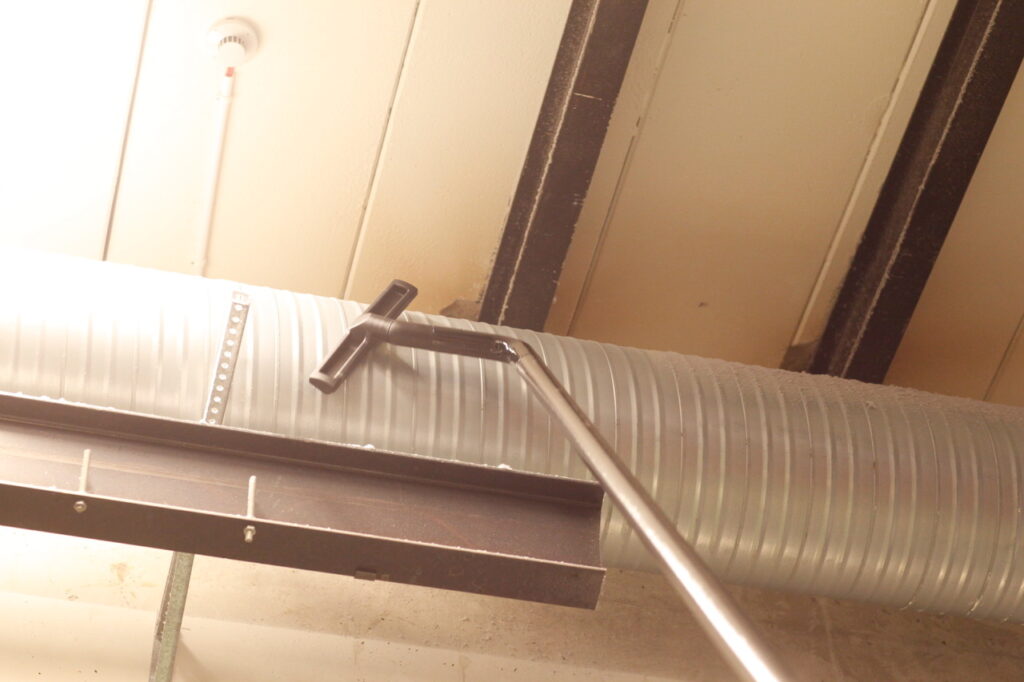 Soft brush head on a carbon-fibre pole cleaning dust from a steel beam beside ducting