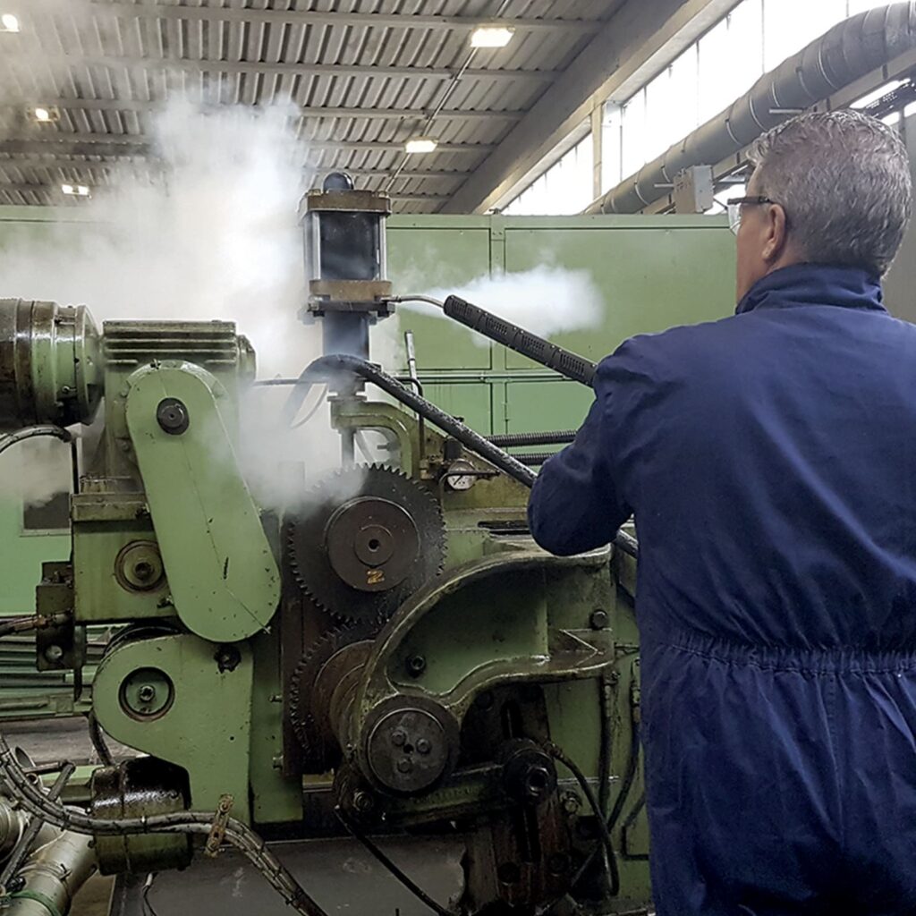 Operator dry-steam cleaning a heavy machine, the same degreasing method used in olive oil mill cleaning