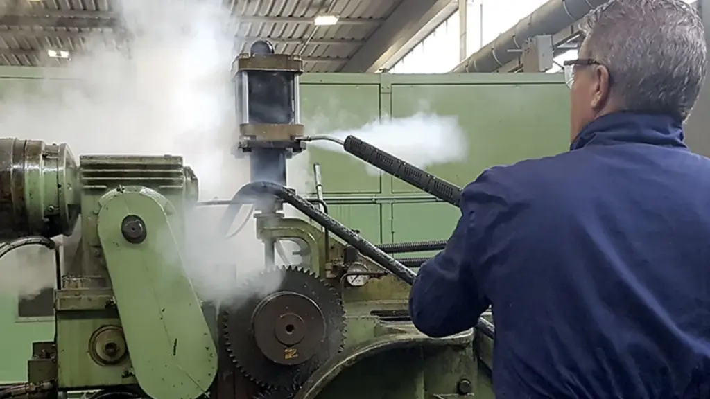 Operator using dry steam degreasing on industrial machinery, with a steam cloud rising from a heavy machine tool