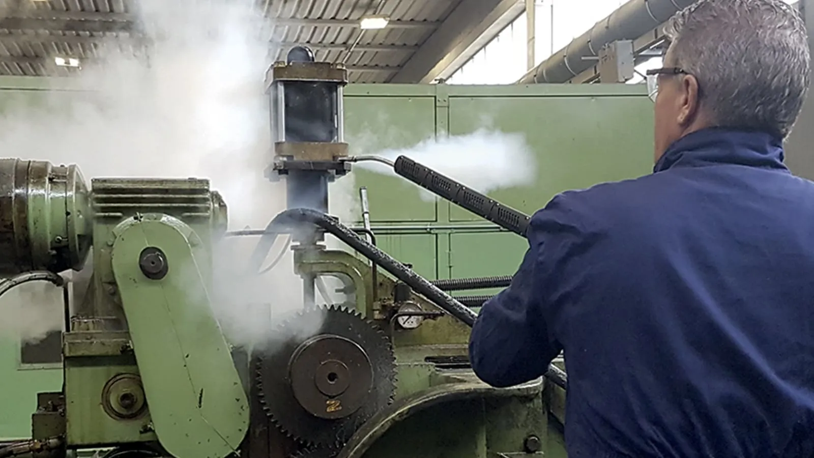 Operator using dry steam degreasing on industrial machinery, with a steam cloud rising from a heavy machine tool
