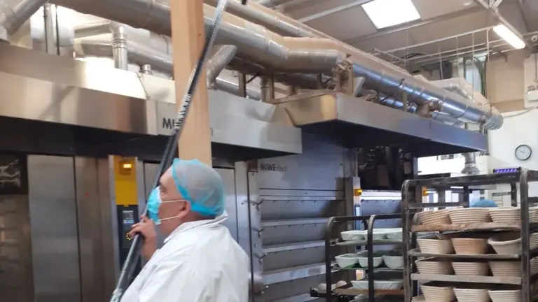 Operator in white coat and hairnet cleaning at height in a large production kitchen
