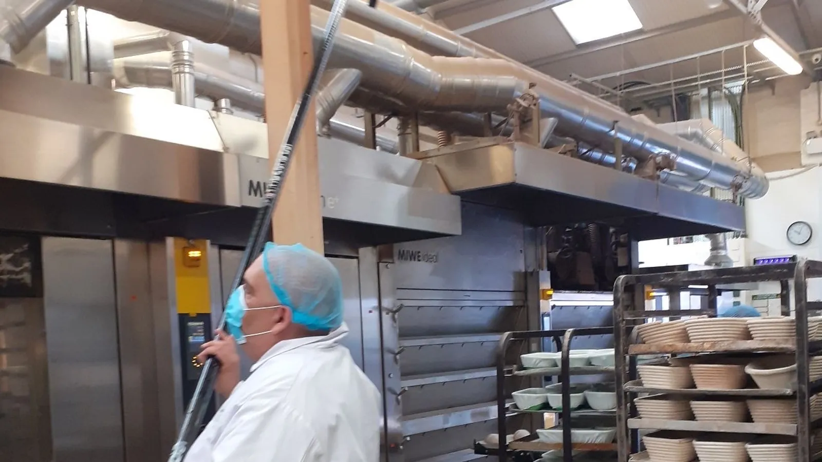 Operator in white coat and hairnet cleaning at height in a large production kitchen
